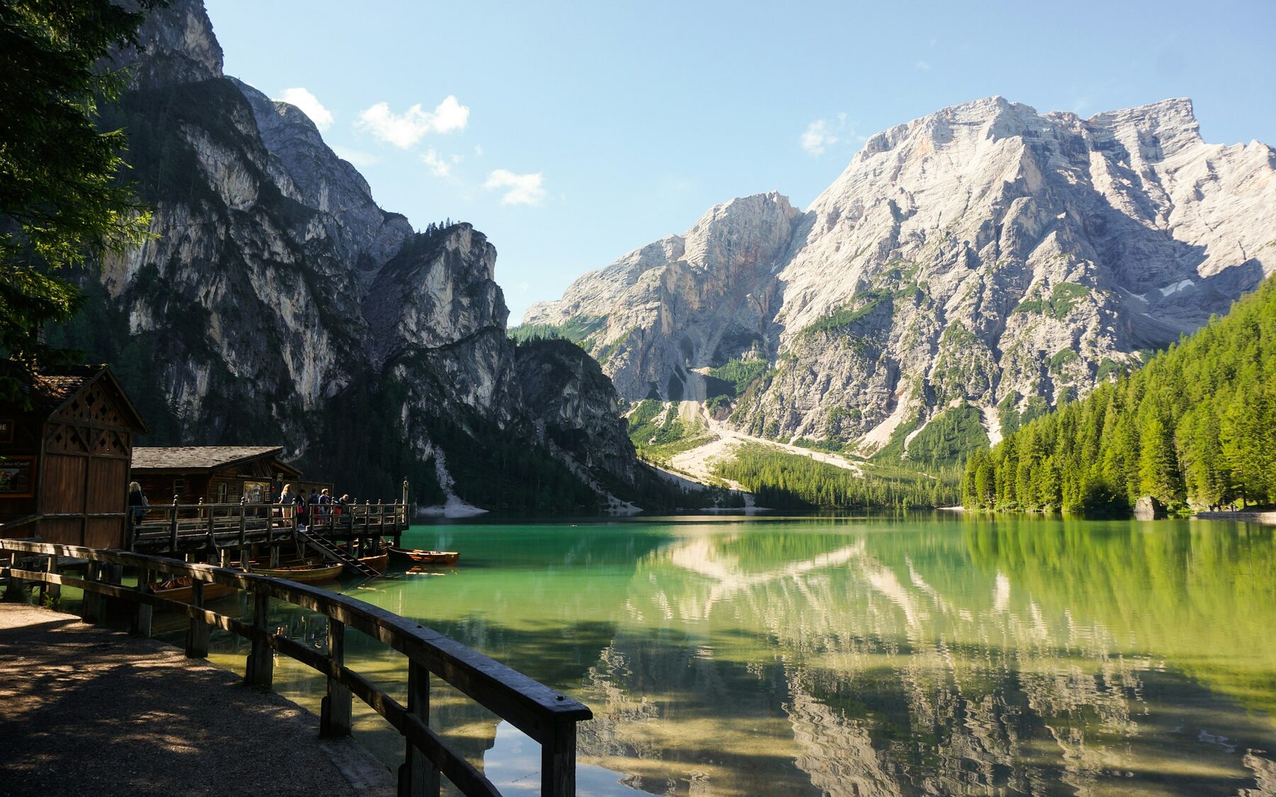Un lago verde y quieto con la pared de la montaña reflejada entera, y a un lado una caseta de madera con barcas amarradas