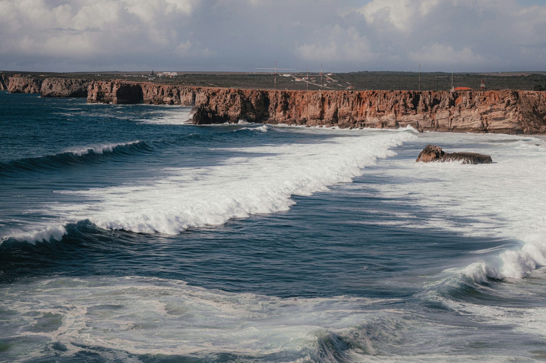Series de olas blancas entrando en línea una detrás de otra hacia una costa baja de acantilado rojizo